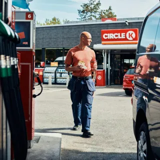 Man walking back to his vehicle at a Circle K station, with a sandwich and coffee in hand